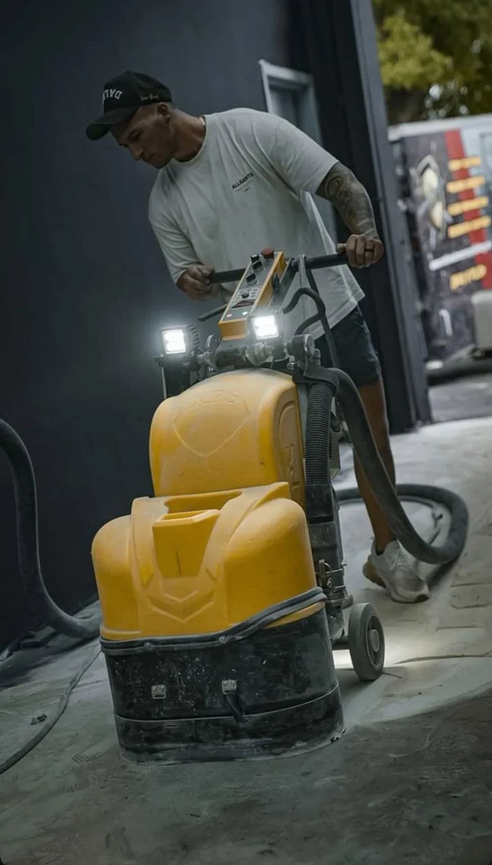 Worker using a concrete grinder on a floor in a modern construction site, with bright lights.