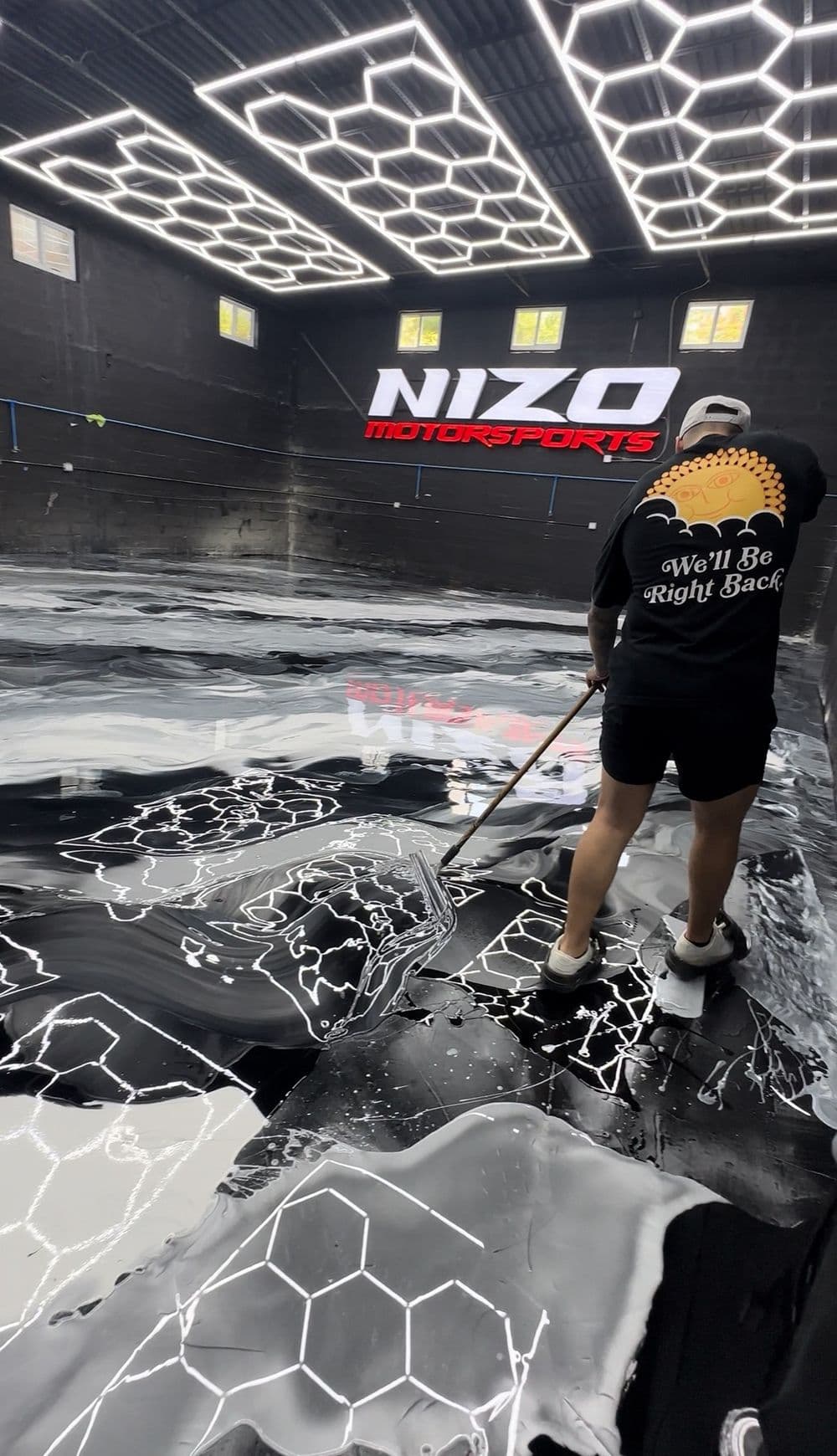 Person applying epoxy flooring at Nizo Motorsports with hexagonal lighting and black surface.