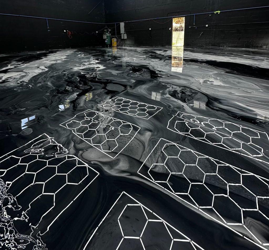 Wide view of a black, gray and white metallic epoxy floor with white hexagon line inlays across a large dark-walled shop interior