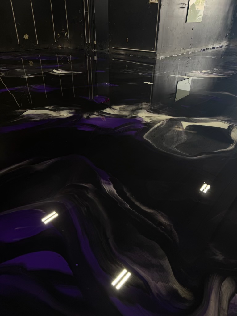 Dark metallic epoxy floor with black, gray and purple swirls reflecting overhead strip lights in a black-walled room