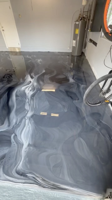 Residential garage with white walls, a water heater and a bicycle, floor coated in a gray, white and charcoal metallic epoxy with flowing swirls